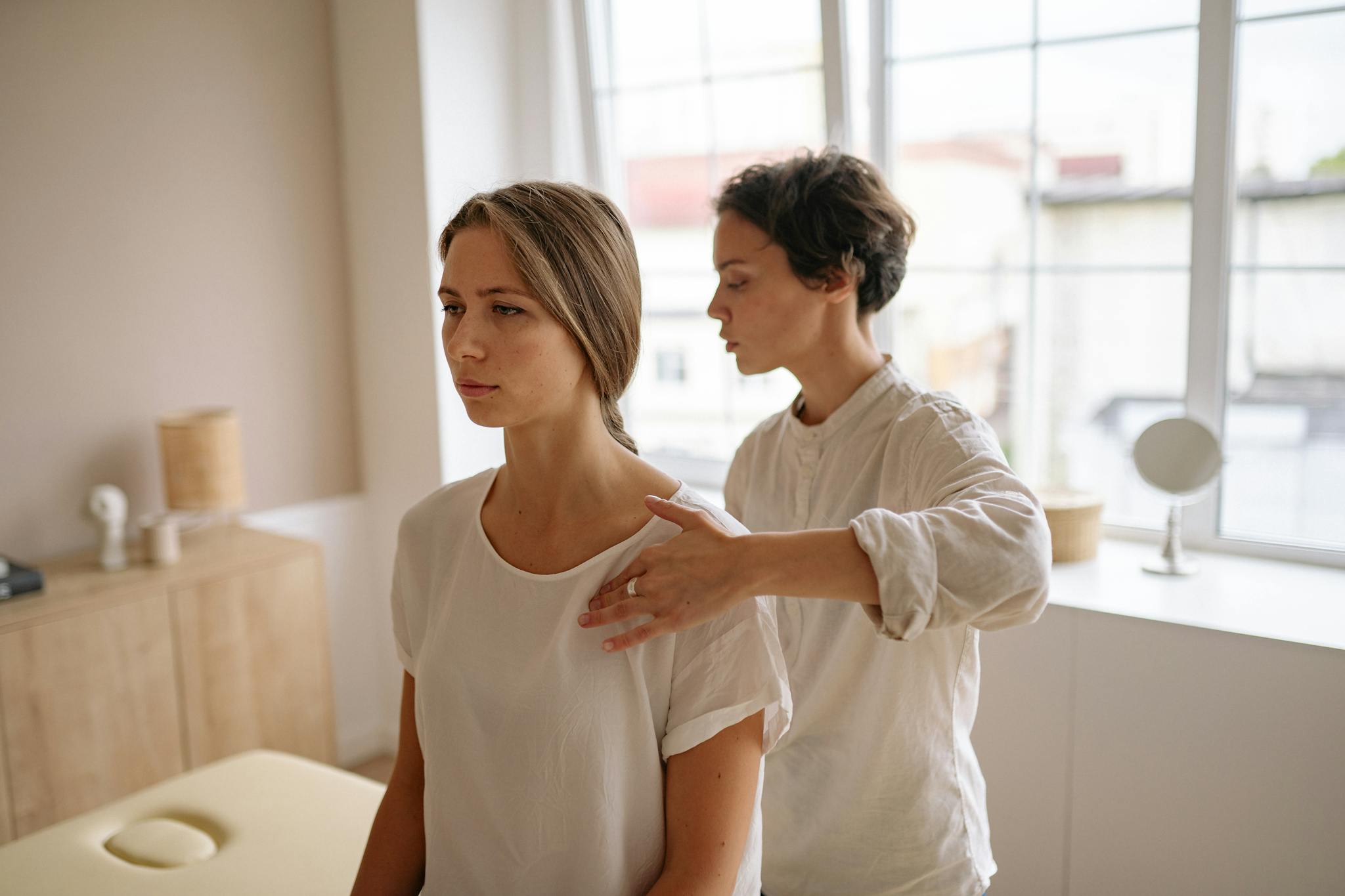 A woman receives shoulder therapy from a therapist in a serene room with natural light.